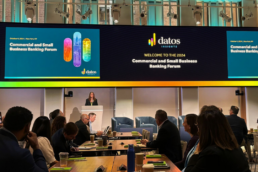 The inside of the Datos Insights Conference, a large colorful screen with the conference title on it while people observe someone presenting.